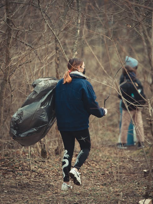 In a dense forested area, there is a collection of discarded waste materials scattered across the leaf-covered ground. Prominently, a large, crumpled blue plastic garbage bag is partially open, reveal