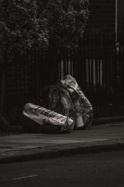A large, weathered and stained beige bulk shopping bag made of heavy-duty plastic or woven material is positioned on a narrow, inclined urban street pavement near a stone or concrete building wall. Th